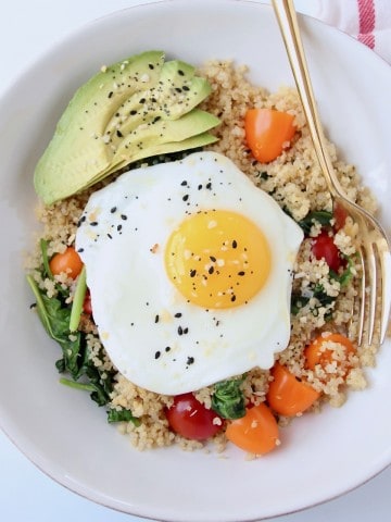 Overhead image of sunny side up egg in bowl with gold fork, sliced avocado and tomatoes