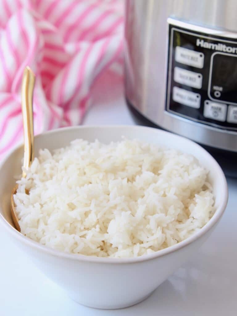 white rice in bowl with gold spoon in front of rice cooker