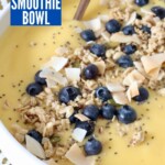 mango smoothie in bowl with spoon and granola, coconut and blueberries on top